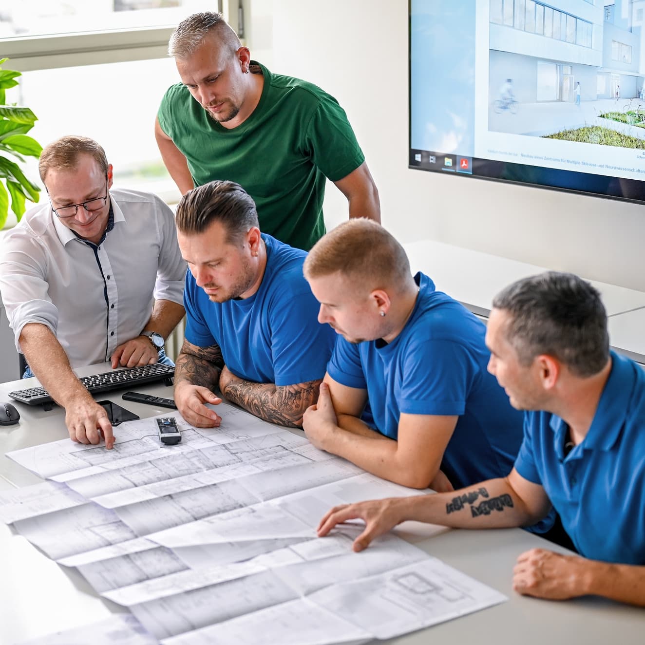 Fünf Mitarbeiter aus dem Bereich Bauen betrachten während einer Besprechung zwei Baupläne