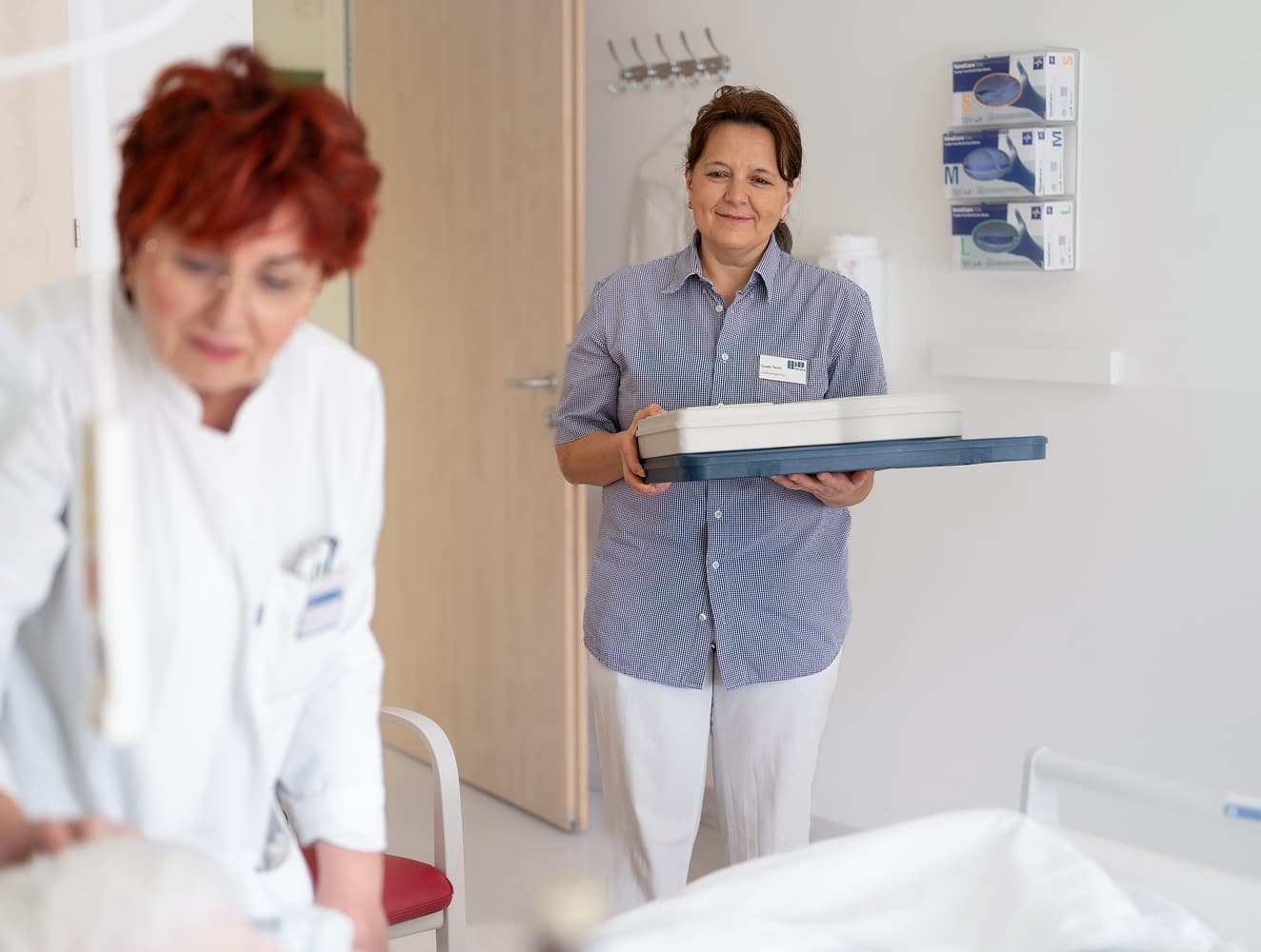 Eine Mitarbeiterin aus dem Bereich Service bringt das Patientenessen ins Patientenzimmer, während eine Ärztin noch im Gespräch mit dem Patienten ist