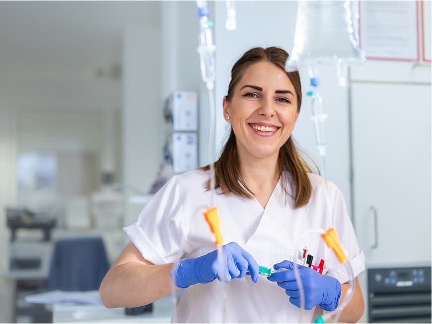 Eine Medizinische Fachangestellte hält eine Infusion in der Hand und blickt in die Kamera